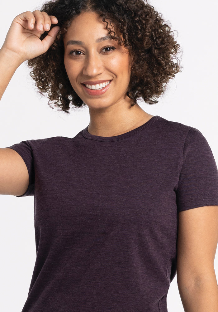 A person with curly hair smiles warmly, raising one hand near their head. They are wearing a lightweight Liza Crew Neck Tee in Deep Plum from Woolx, and they stand against a plain white background.