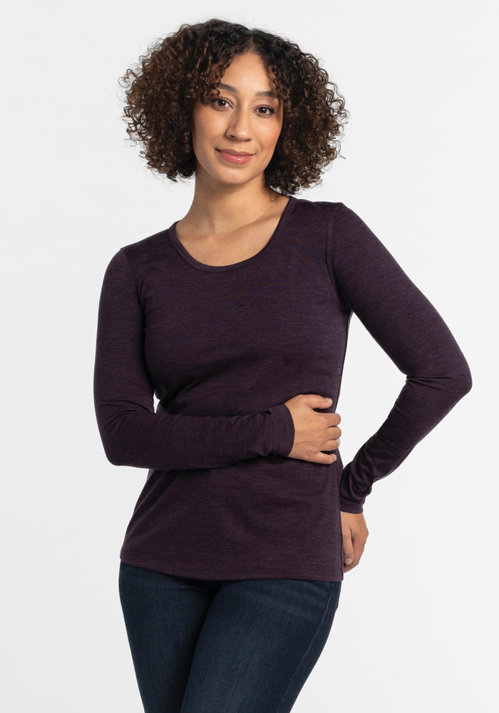 A person with curly hair is wearing the Remi Long Sleeve T-Shirt in Deep Plum by Woolx and a pair of jeans. They stand against a plain white background, smiling gently with their arms relaxed, enjoying the comfortable fit of the lightweight fabric. 