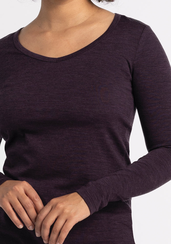 A person is standing against a plain white background, wearing a Woolx Layla V Neck Top in Deep Plum. Their head is not visible, and their hands are gently clasped in front of them.