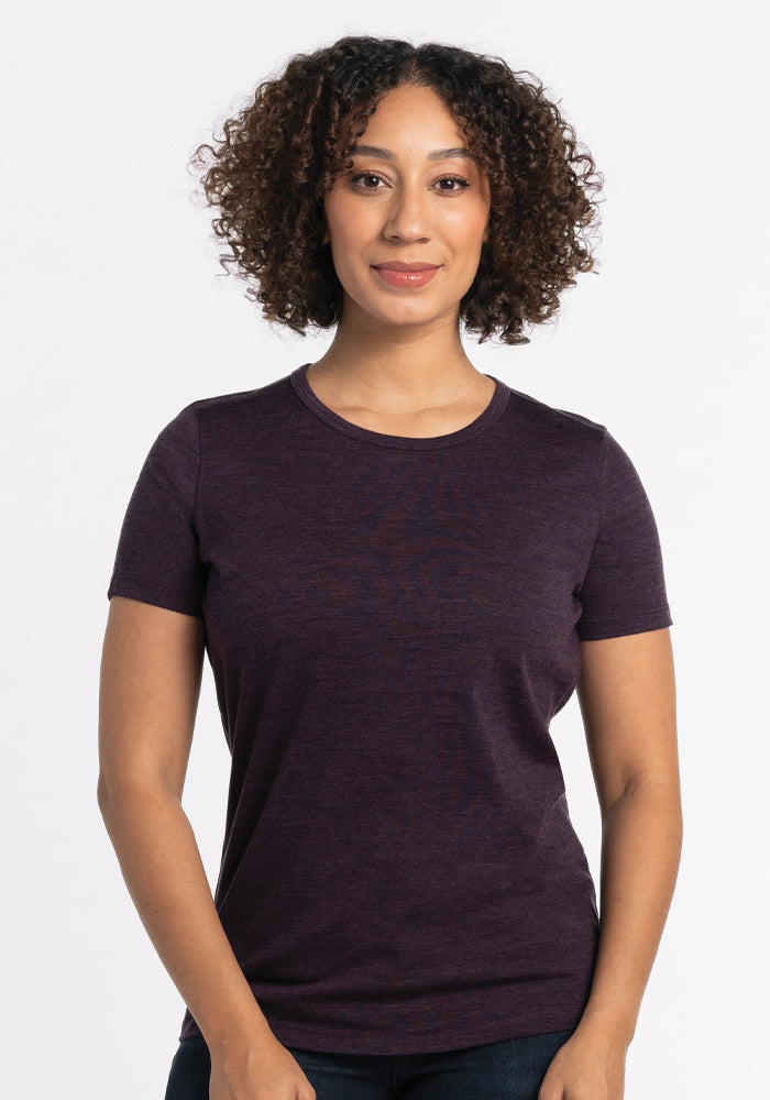 A person with curly hair is wearing a Woolx Liza Crew Neck Tee in Deep Plum, crafted from lightweight fabric, along with jeans. They stand against a white background, displaying a neutral expression and facing forward.