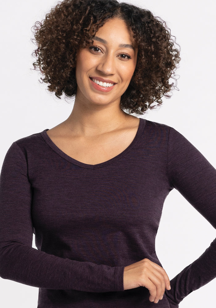 A person with curly hair smiles at the camera, wearing the Woolx Layla V Neck Top in Deep Plum, made from lightweight fabric. The background is plain white.