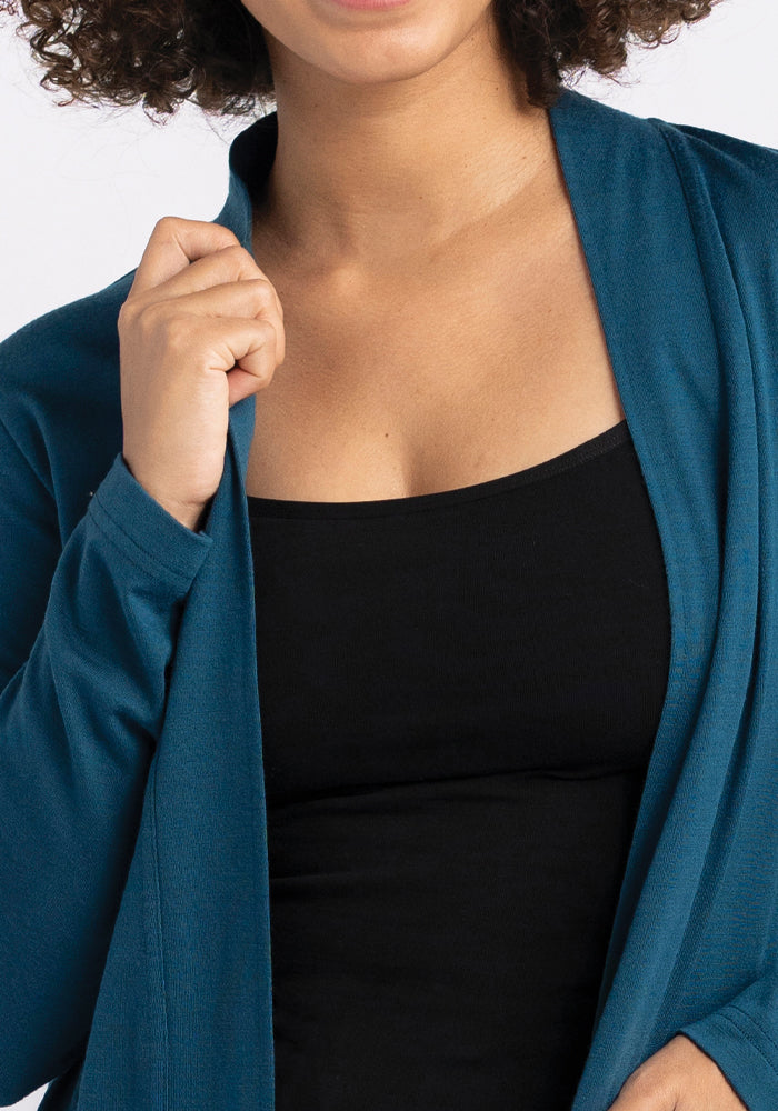 A person with curly hair is wearing a black top and the "Paisley Cardigan - Real Teal" by Woolx, crafted from a Duralite blend. They are holding the front of the cardigan with one hand, showcasing their upper torso and part of their face.