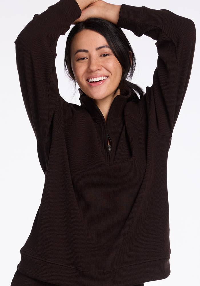 A person with long dark hair is smiling with their arms raised above their head, wearing a sleek travel outfit featuring the Woolx Quinn Ribbed 1/4 Zip in French Roast. The plain white background highlights their joyful expression and stylish ensemble.