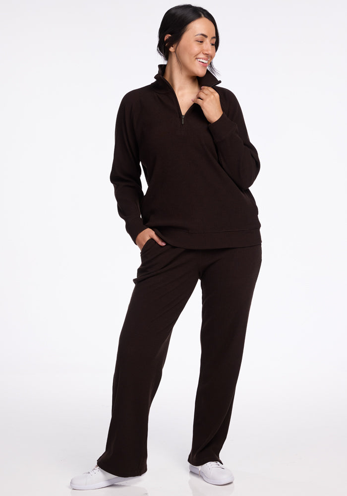 A person with long dark hair smiles and looks to the side. They are wearing a Woolx Quinn 1/4 Zip merino wool long-sleeve top in a dark color, paired with matching pants, and white sneakers. Their left hand is in their pocket, while their right hand rests near their chin—a perfect addition to any fall wardrobe.