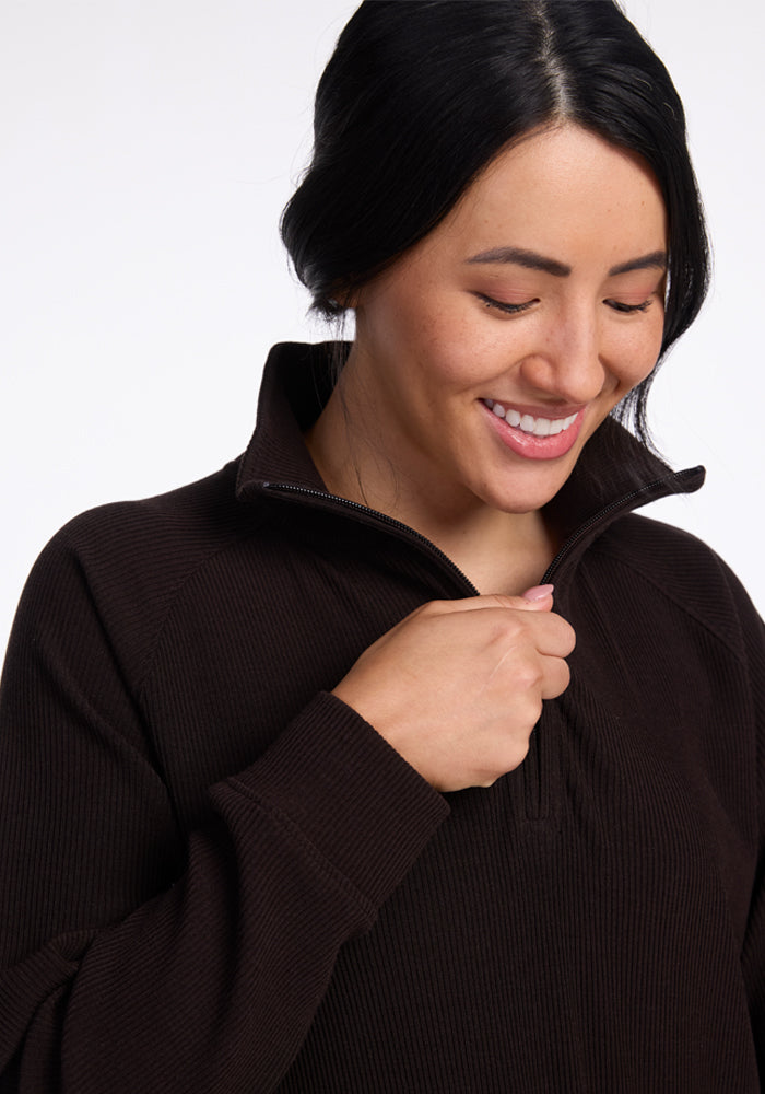 A person with long dark hair is smiling and looking downwards while holding the zipper of their Woolx Quinn 1/4 Zip, ribbed sweater. The merino wool garment is partially unzipped at the collar. The background is plain and white, highlighting the person and their cozy fall wardrobe. - French Roast