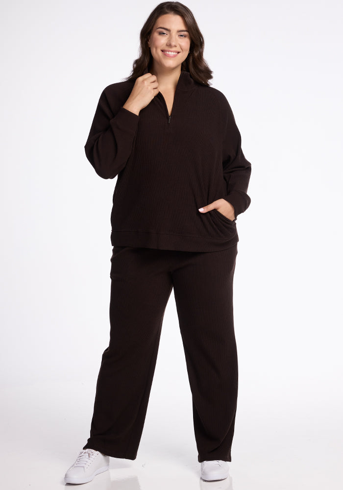 A woman stands smiling against a white background. She is wearing the black Quinn 1/4 Zip Merino wool top from Woolx's fall ribbed collection along with matching black pants. Her left hand is in her pocket, and she is sporting white sneakers. A cozy stylish layer perfect for any occasion.