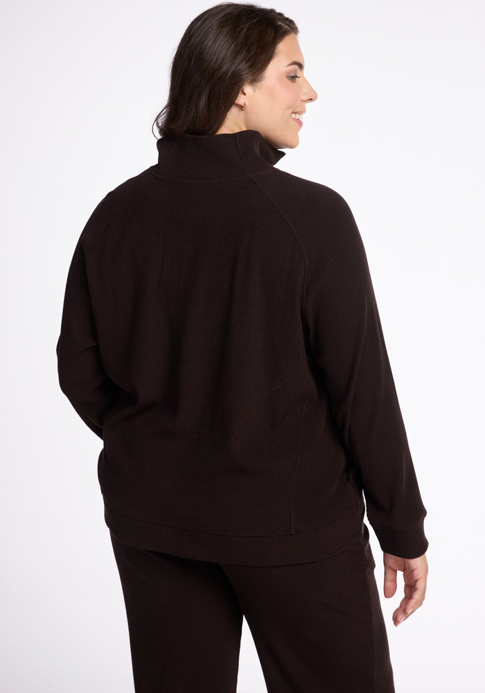 A person with long, dark hair is standing and facing away from the camera, wearing a stylish Quinn 1/4 Zip top and matching dark pants from Woolx’s fall ribbed collection. The backdrop is plain white.