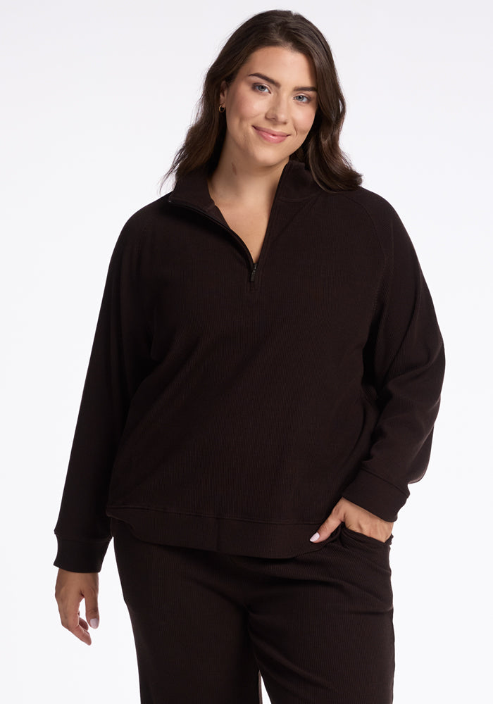 A person with long dark hair is posing against a white background, smiling slightly. They are wearing a black Woolx Merino wool Quinn 1/4 Zip shirt from the fall ribbed collection and matching pants. One hand rests casually in their pocket. 