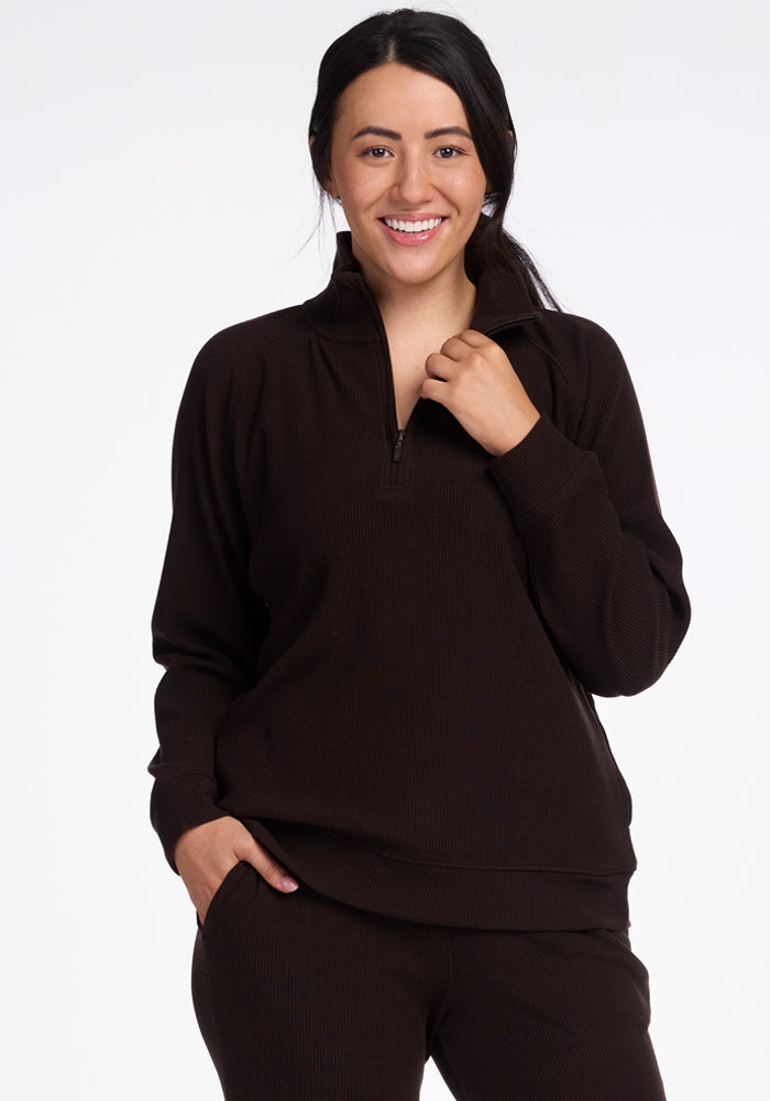 A person with long dark hair smiles at the camera, wearing a dark brown Woolx Quinn 1/4 Zip sweater and matching pants. They have one hand in their pocket and the other touching the collar of their cozy merino wool sweater. The background is plain white, showcasing a perfect fall wardrobe. 