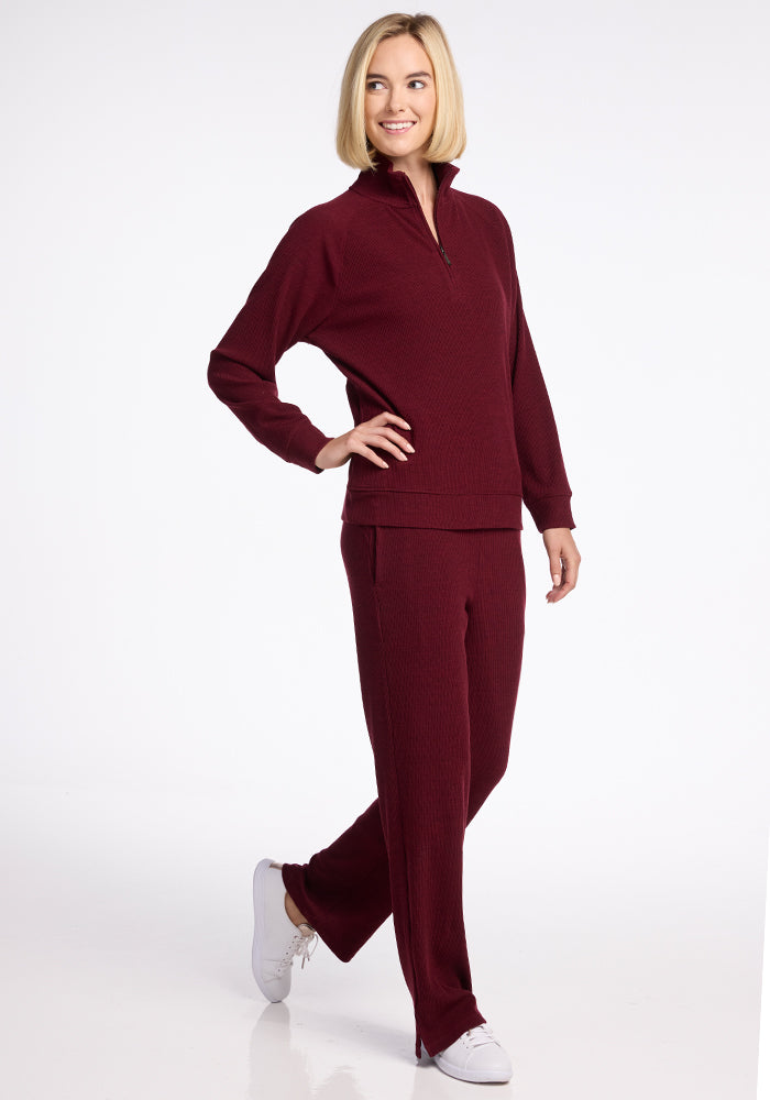 A person with short blonde hair, smiling, is wearing a maroon long-sleeve Woolx Quinn 1/4 Zip top with matching maroon pants and white sneakers. They are standing in a relaxed pose against a plain white background.