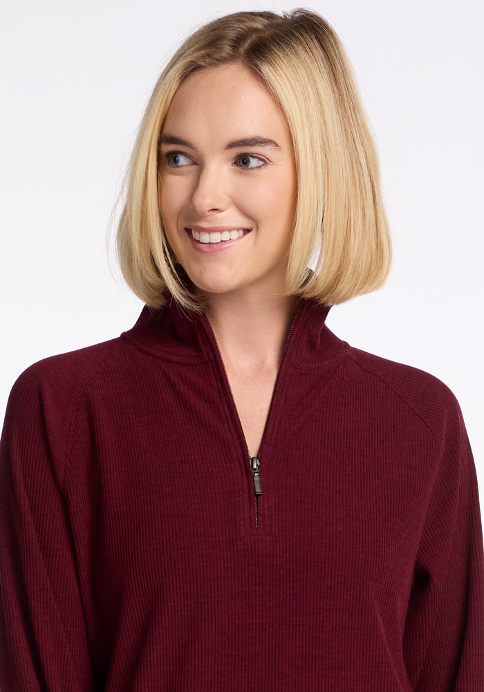 A person with shoulder-length blonde hair smiles while looking off to the side, wearing the Woolx Quinn Ribbed 1/4 Zip in Cranberry Melange. The background is a neutral, light color.