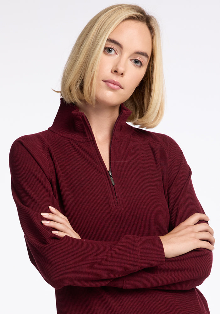 A person with straight blonde hair and a neutral expression poses with arms crossed, wearing a long-sleeved, burgundy Quinn 1/4 Zip top from Woolx, made from Merino wool. The high collar enhances the sophisticated athleisure quarter zip style. The background is simple white. 