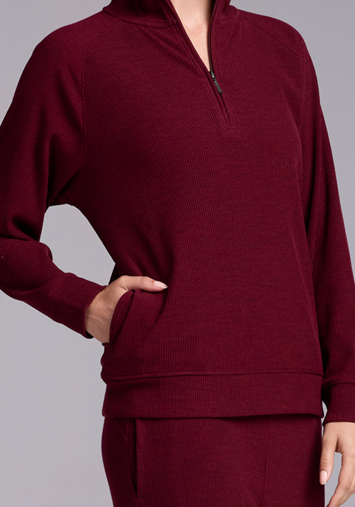 A person is wearing a matching maroon ribbed sweater and pants made from soft Merino wool. The Woolx Quinn 1/4 Zip sweater features a quarter-zip front and a high collar. The person's left hand is in the pocket of the pants. The background is plain and gray.