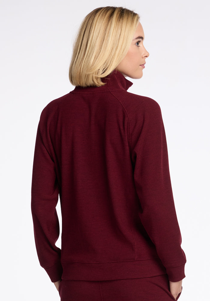 A person with shoulder-length blonde hair is viewed from behind, donning a long-sleeved, high-collar Woolx Quinn 1/4 Zip sweater in burgundy, made of Merino wool. The background is plain white.