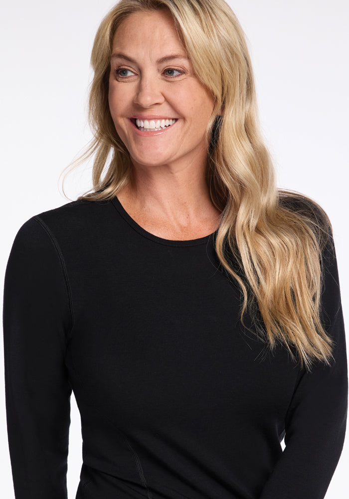 A smiling woman with long blonde hair is wearing the Woolx Riley Long Sleeve Crew in black. She is looking slightly to the side against a neutral background.