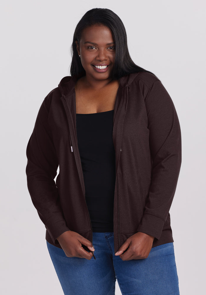 A person with long hair smiles while wearing an athletic fit black top under a Ryann Hoodie in French Roast from Woolx, paired with blue jeans. They stand against a plain white background. 