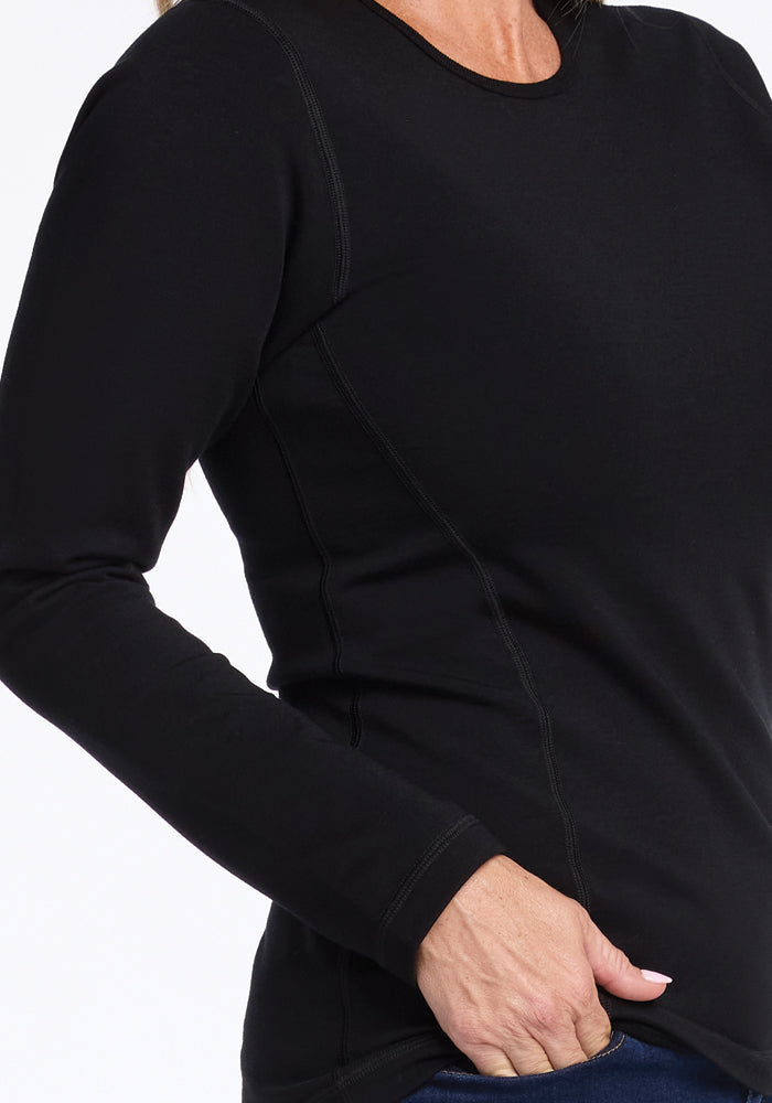 A person wearing the Riley Long Sleeve Crew in black by Woolx stands with their hand in the pocket of dark pants. The focus is on the upper half of this insulating shirt, highlighting its intricate stitching and soft Merino wool texture.