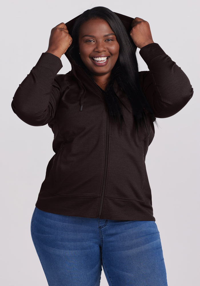 A smiling person wearing the Woolx Ryann Hoodie in French Roast, with the hood up and paired with blue jeans, stands against a plain background.