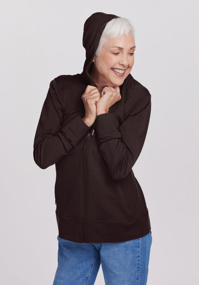 An older person with short white hair smiles while wearing the Woolx Ryann Hoodie in French Roast and blue jeans. They hold the hoodie collar with both hands against a plain gray background.