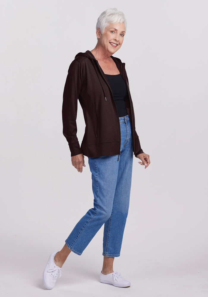 An older woman with short gray hair is smiling and posing. She is wearing a black top, the Woolx Ryann Hoodie in French Roast, blue jeans, and white sneakers. The background is plain white.