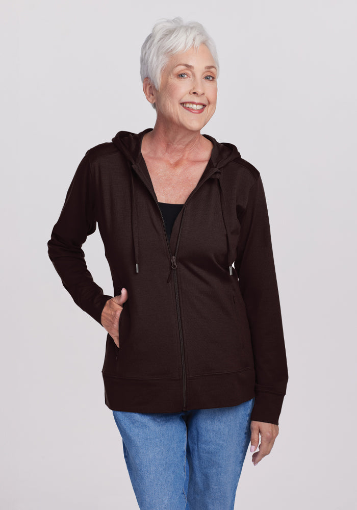 An older woman with short white hair smiles warmly as she wears the Woolx Ryann Hoodie in French Roast over a black top and blue jeans. With one hand in her pocket, she stands against a plain light gray background, exuding comfort and casual style. 