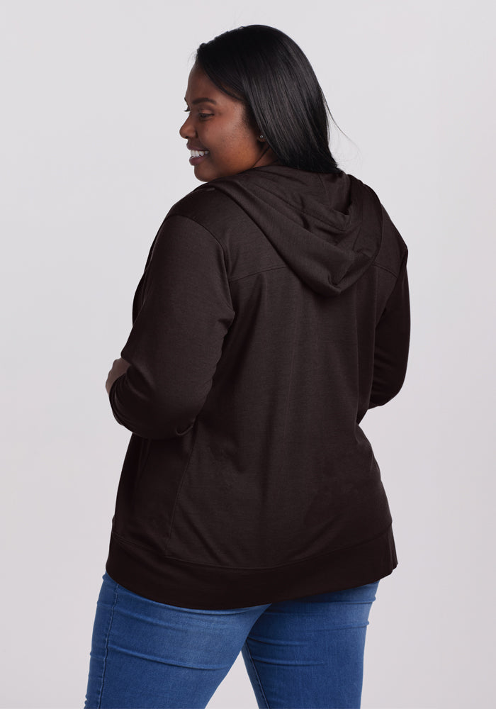 A woman with long black hair is wearing the dark brown Ryann Hoodie in French Roast from Woolx and blue jeans. The athletic fit accentuates her form as she smiles, turned slightly sideways and looking off to the side, against a plain gray background.