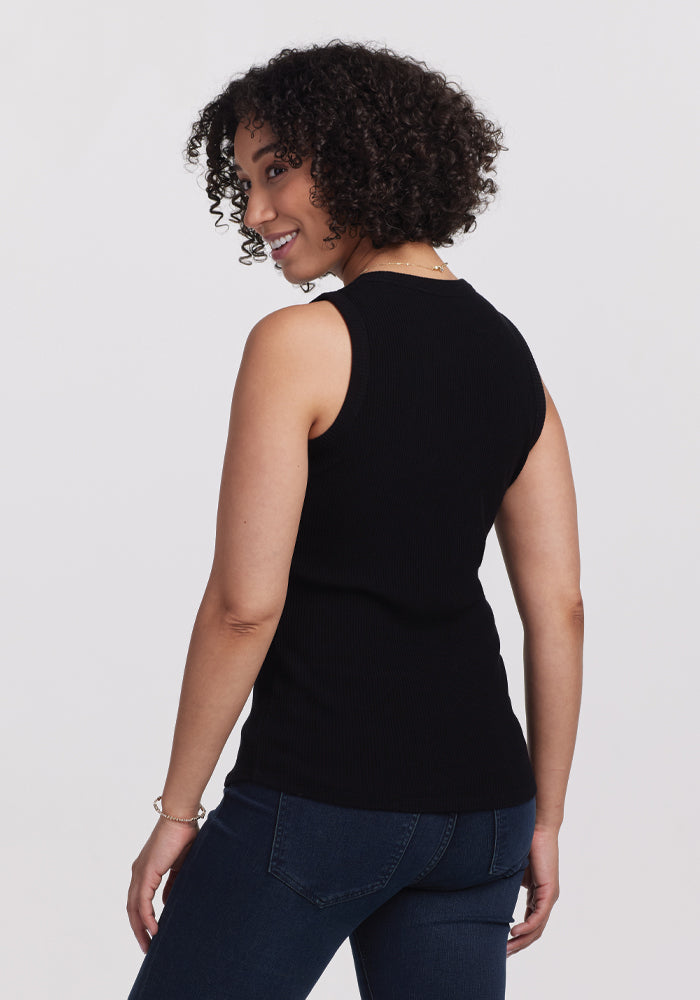 A person with curly hair wears a sleek black Raegan Ribbed Tank Top by Woolx paired with dark jeans, smiling slightly towards the camera against a plain white background.