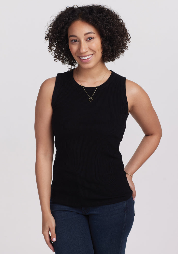 A person with curly hair wearing a Woolx Raegan Ribbed Tank Top in black and blue jeans smiles while standing against a plain background. 