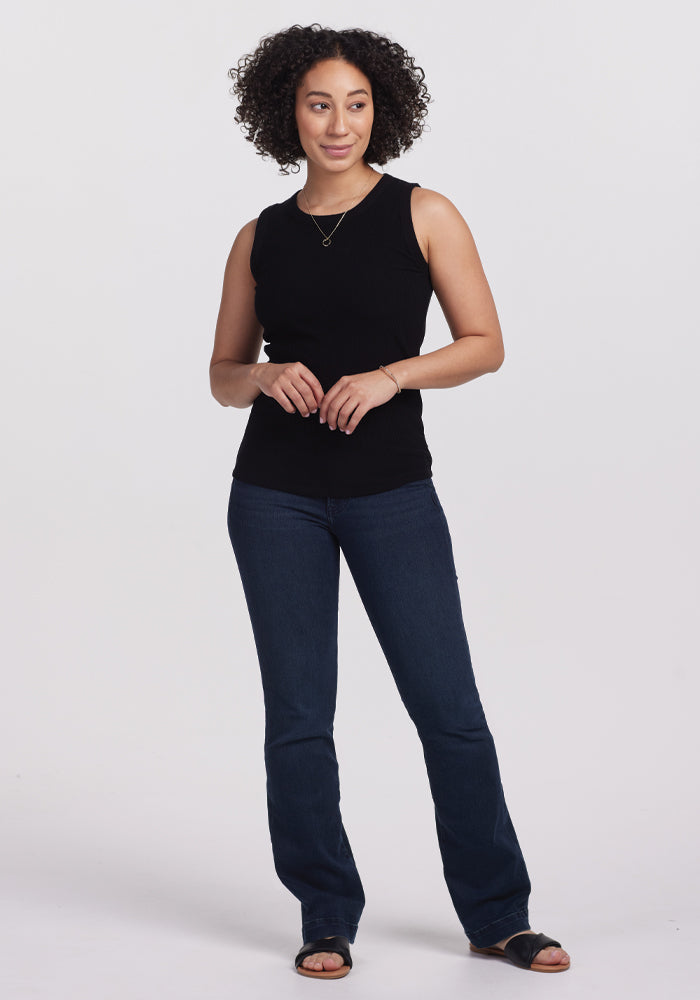 A person with curly hair poses against a plain background, wearing a black Raegan Ribbed Tank Top from Woolx, blue jeans, black sandals, and a simple necklace. They stand relaxed with gently clasped hands.