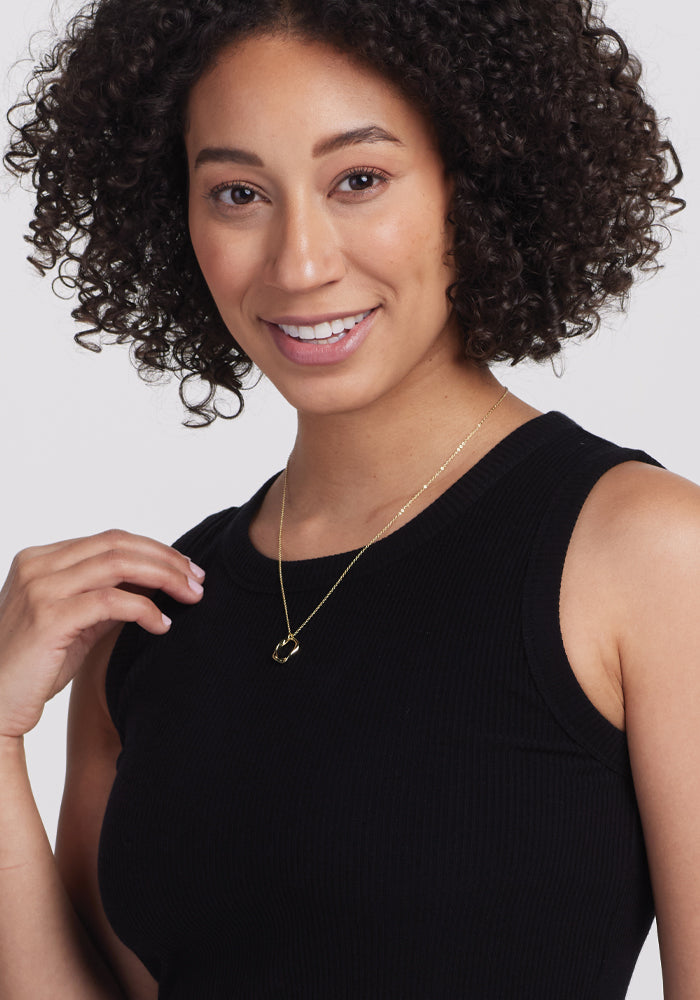 A woman with curly hair smiles at the camera, wearing a Woolx Raegan Ribbed Tank Top in black and a gold pendant necklace. She gently touches the necklace with one hand against a plain light background.