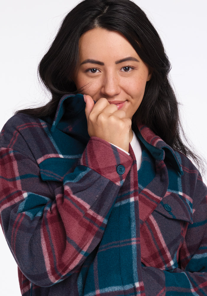 A person with long dark hair partially covers their mouth with the collar of their plaid Sawyer Shacket by Woolx, which features dark blue, maroon, and grey colors. They are looking slightly off-camera, giving a soft and thoughtful expression.