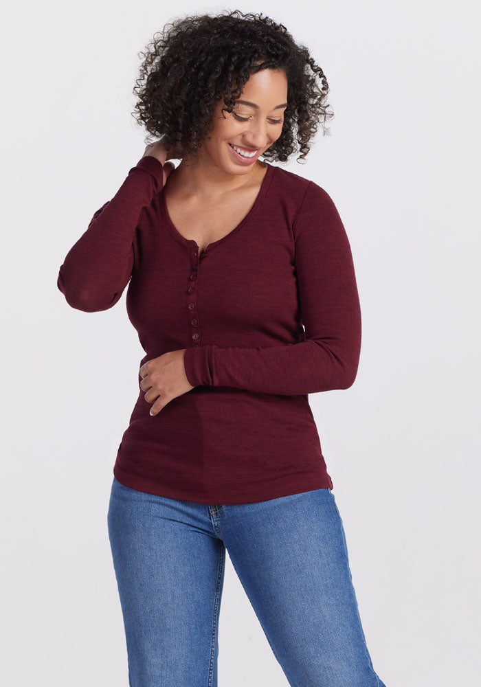 A person with curly hair smiles while looking down and to the side, touching the back of their neck with one hand and their waist with the other. They are wearing a Woolx Reese Ribbed Top in Cranberry Melange and blue jeans against a plain white background. 