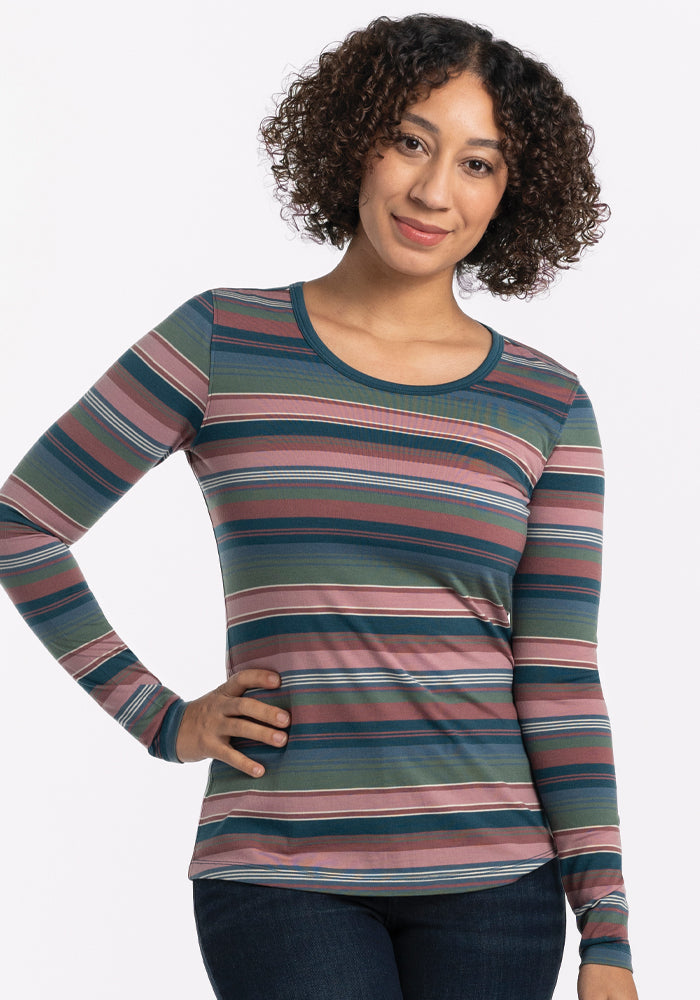 A person with curly hair smiles while wearing the Woolx Remi Long Sleeve T-Shirt in Adirondack 2.0 Stripe, made of lightweight fabric. Their hand rests on their hip against the plain white background. 