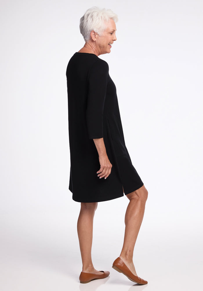 An older adult with short white hair is wearing the Woolx Rissa A Line Dress in black along with brown flat shoes. They are standing in profile view against a plain white background, highlighting the effortless elegance of their attire.