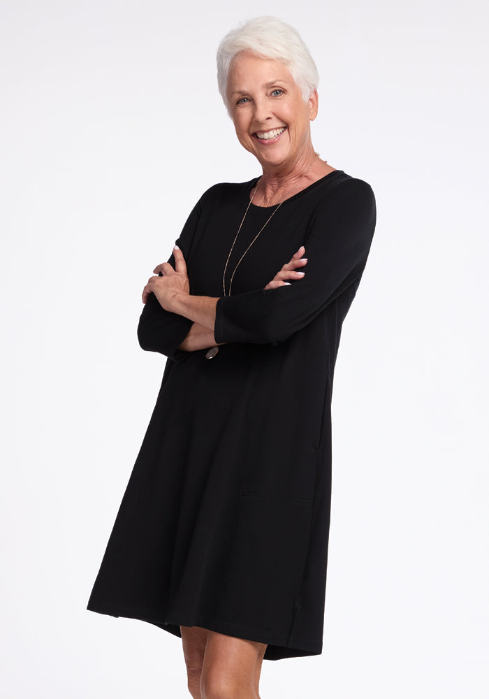 A woman with short white hair smiles confidently, arms crossed in the layer-friendly Rissa A Line Dress from Woolx. Her black outfit is complemented by a long necklace, all against a plain white backdrop.