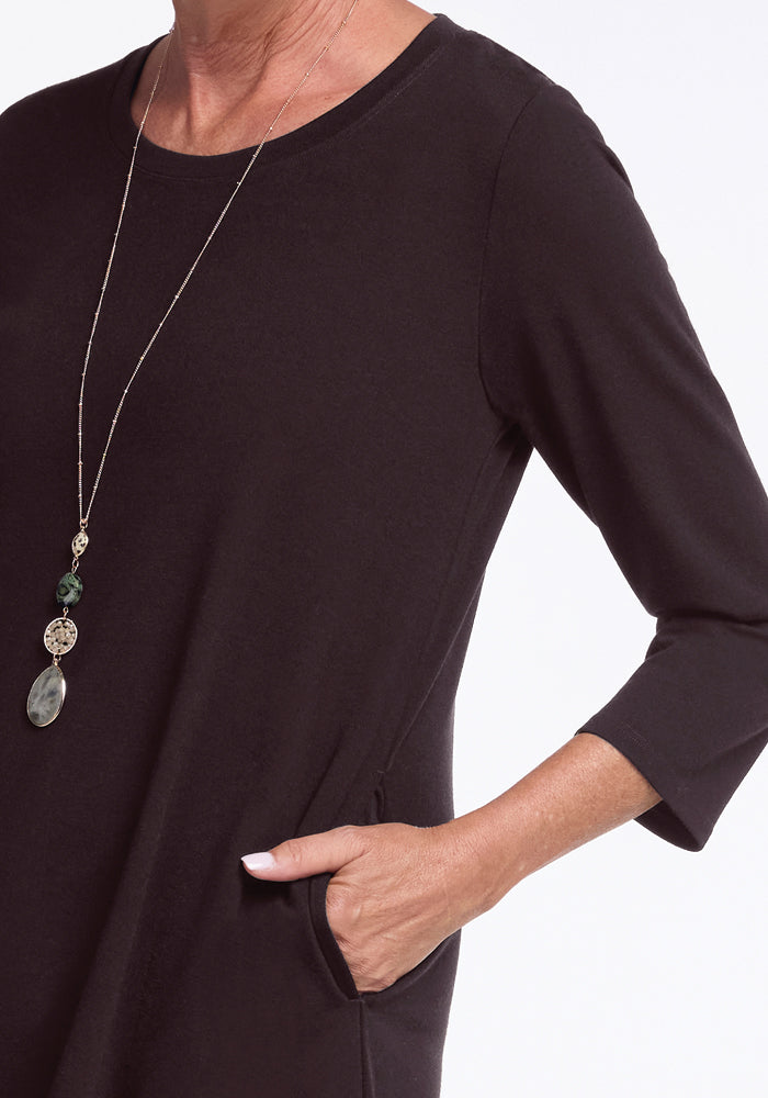 Styled elegantly, the individual dons the Rissa A Line Dress in French Roast by Woolx. The refined touch of Merino Wool fabric enhances the look, accompanied by a long necklace with decorative pendants. The image captures one hand resting in a pocket, focusing on the upper torso and arm against a white background.