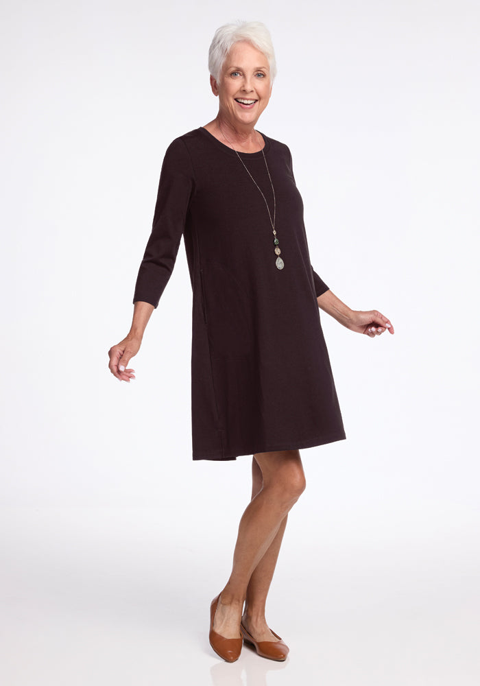 A woman with short white hair is smiling while posing in a chic Rissa A-Line Dress - French Roast by Woolx, paired with brown shoes. Her ensemble is accentuated by a long necklace, arms slightly outstretched, set against a plain white studio backdrop.