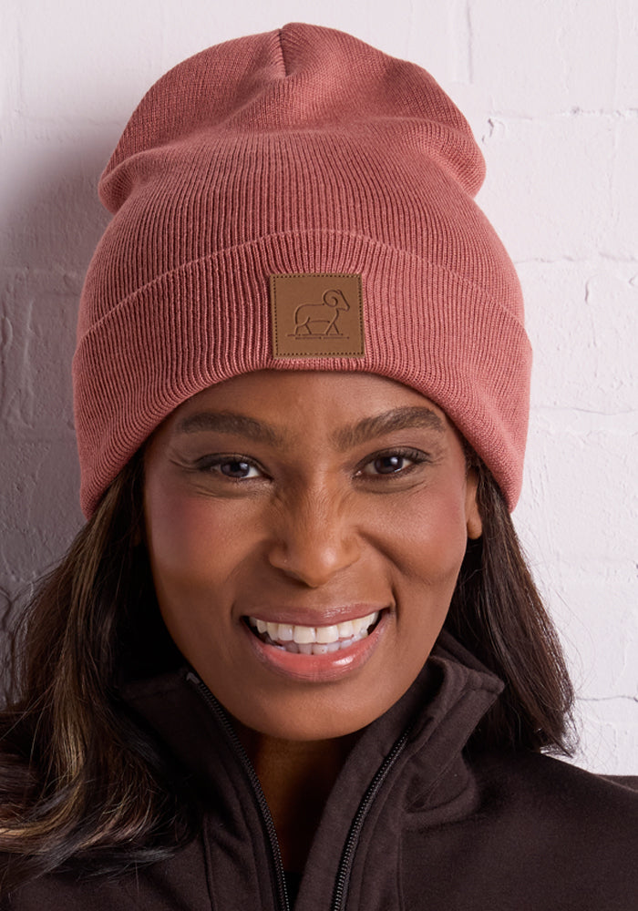 A woman smiles while wearing the Baylor Beanie - Brick Dust from Woolx, a cute knit design featuring a leather patch logo on the front. She's dressed in a dark jacket and stands against a white brick wall, effortlessly combining style and warmth.