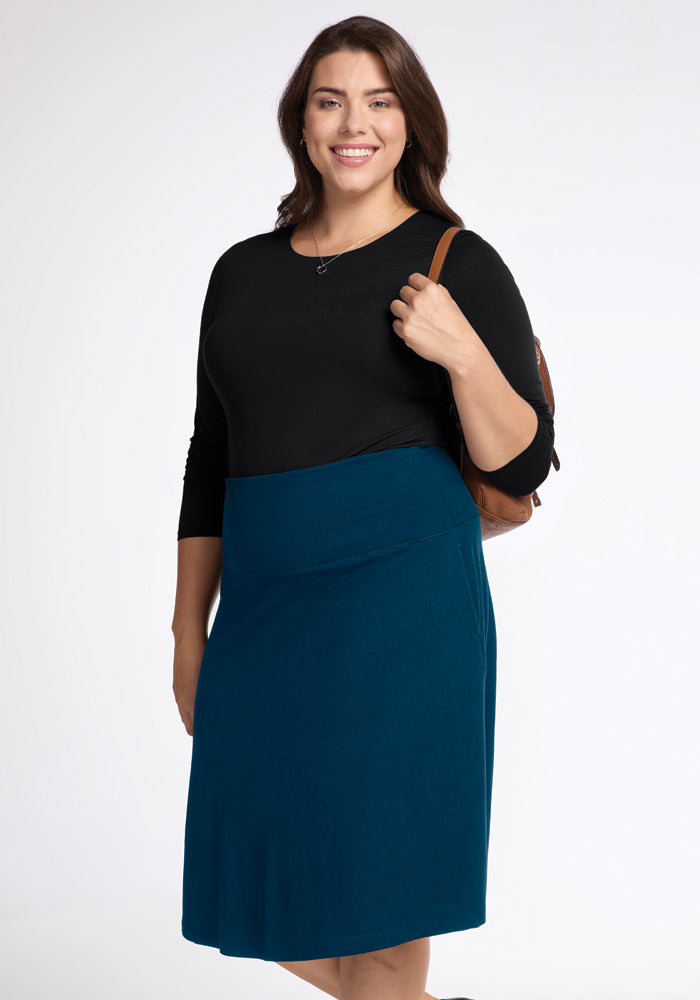 A person with long brown hair is smiling, wearing a black top and a Marilyn Flowy Midi Skirt in Real Teal by Woolx. They have a brown backpack slung over one shoulder and are standing against a plain white background.