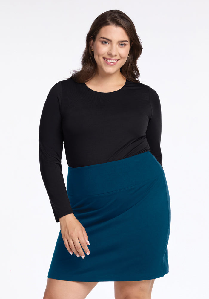 A woman with long dark hair is smiling and posing in a travel-friendly ensemble. She wears a black long-sleeve top paired with the Woolx Monroe Knee Length Skirt in Real Teal, which provides moisture-wicking comfort. The background is plain white.