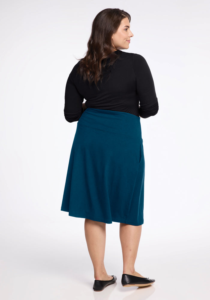 A woman with long dark hair stands with her back turned, wearing a black long-sleeve top and the Marilyn Flowy Midi Skirt in Real Teal by Woolx, featuring an A-Line design. Her outfit is completed with black flat shoes against a backdrop of plain light gray.