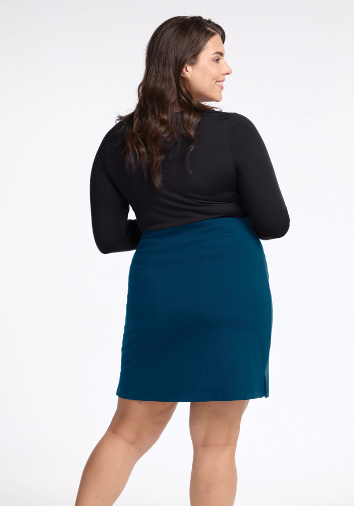 A woman stands with her long wavy brown hair cascading over a black top and the Monroe Knee Length Skirt in Real Teal by Woolx. This travel-friendly, moisture-wicking merino wool skirt is ideal for any journey, while the light-colored background adds simplicity to the scene.