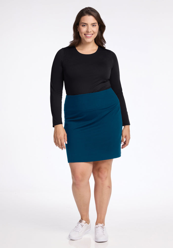 A woman with brown hair stands smiling, wearing a black long-sleeve top and the Monroe Knee Length Skirt in Real Teal from Woolx. She pairs her stylish travel-friendly Merino wool skirt with white sneakers, standing against a plain white background that highlights her comfortable yet chic outfit, ideal for any journey.