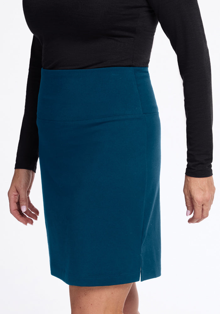 A person stands against a white background, wearing a black long-sleeve top and the Woolx Monroe Knee Length Skirt in Real Teal, expertly crafted from moisture-wicking Merino wool for perfect travel-friendliness.