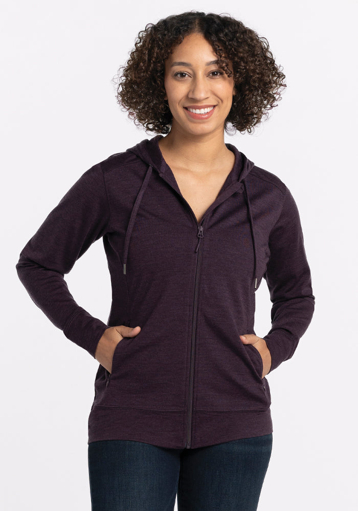 A person with curly hair smiles while wearing the Woolx Ryann Hoodie in Deep Plum, featuring an athletic fit, with hands tucked in the pockets. The background is plain white.