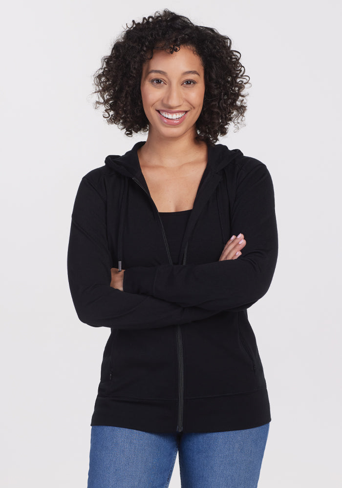 A person with curly hair stands against a white background, arms crossed casually, wearing a Woolx Ryann Hoodie in black paired with blue jeans, perfectly blending comfort and style. 