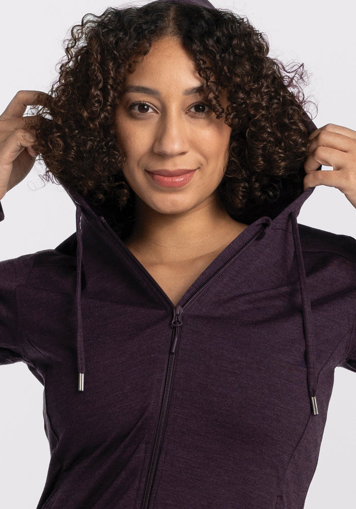 A person with curly hair smiles as they put on the Woolx Ryann Hoodie in Deep Plum, featuring a lightweight and athletic fit. Equipped with zippers and drawstrings, this hoodie is ideal for any adventure. The background is plain and light-colored.