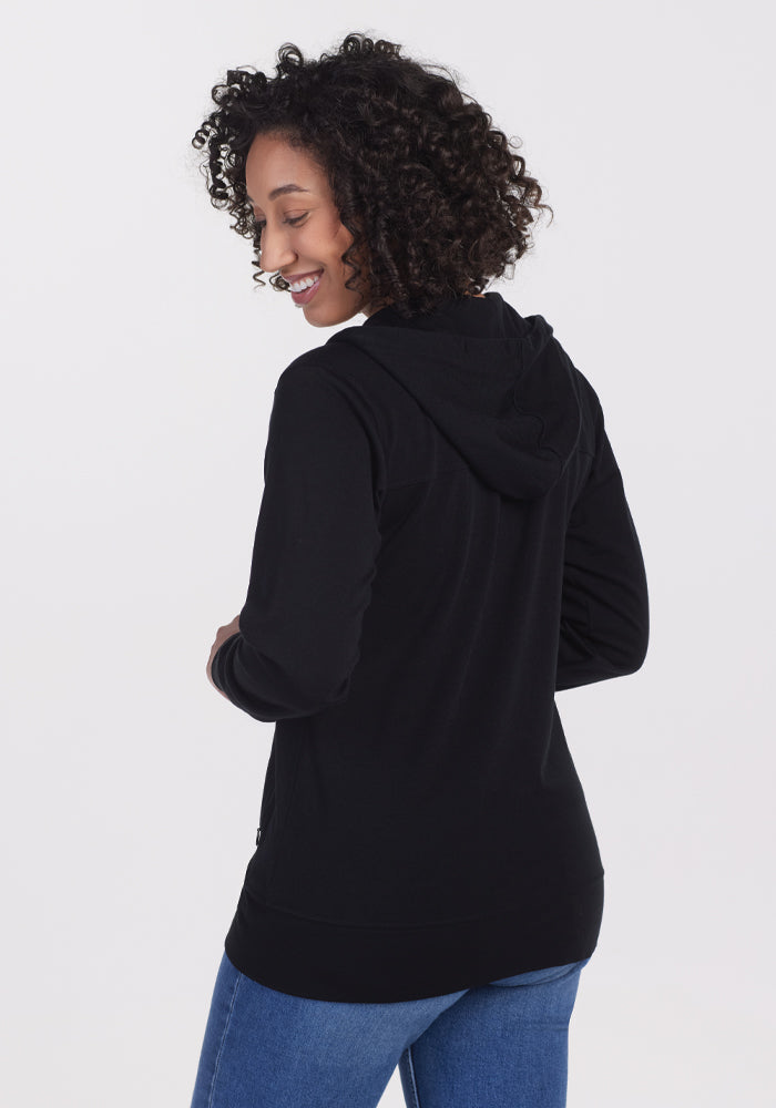 A person with curly hair smiles over their shoulder against a white background, wearing Woolx's black Ryann Hoodie and blue jeans. This lightweight hoodie is ideal for travel essentials, providing all-day comfort.