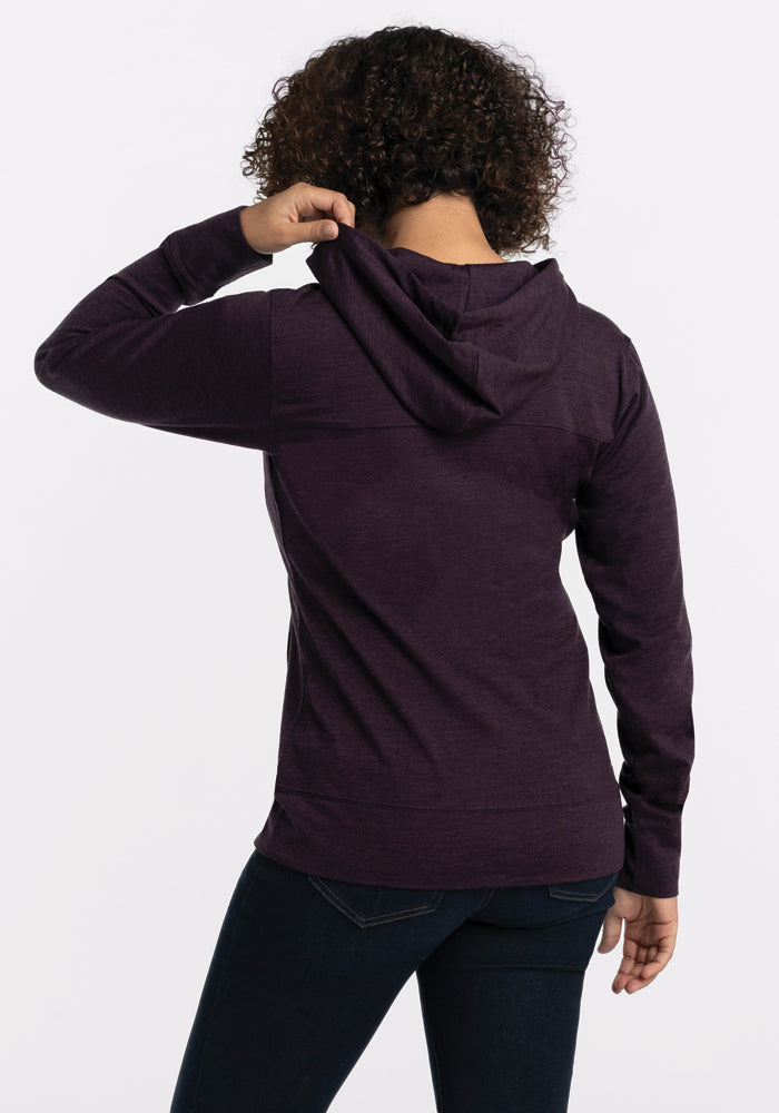 A person with curly hair wears the Woolx Ryann Hoodie in Deep Plum, crafted from lightweight Merino wool. They are standing with their back to the camera, slightly turning their head over their left shoulder while holding the hood with their left hand. The background is plain white.