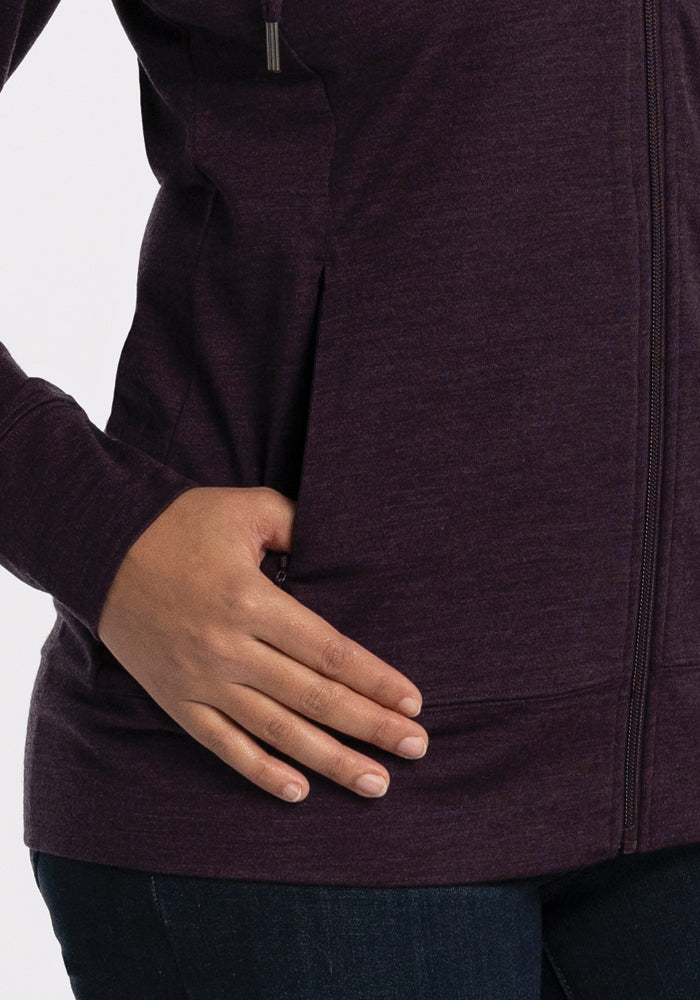 A person with their left hand tucked into the pocket of a Woolx Ryann Hoodie in Deep Plum. The garment, featuring an athletic fit and a visible front zipper, is ideally paired with dark blue jeans.